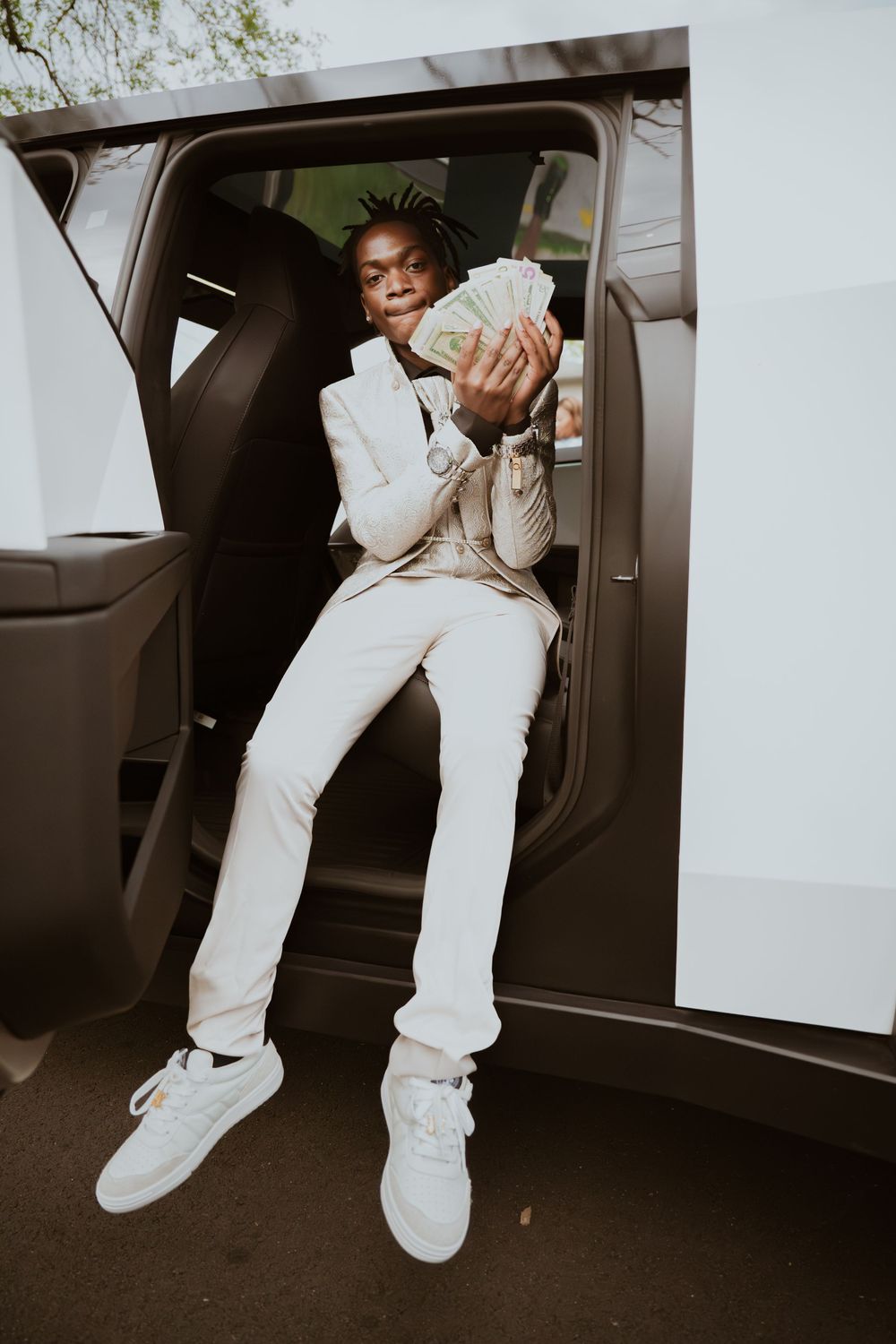 Young man in white suit sitting in car door holding fan of cash bills, stylish urban fashion photo.