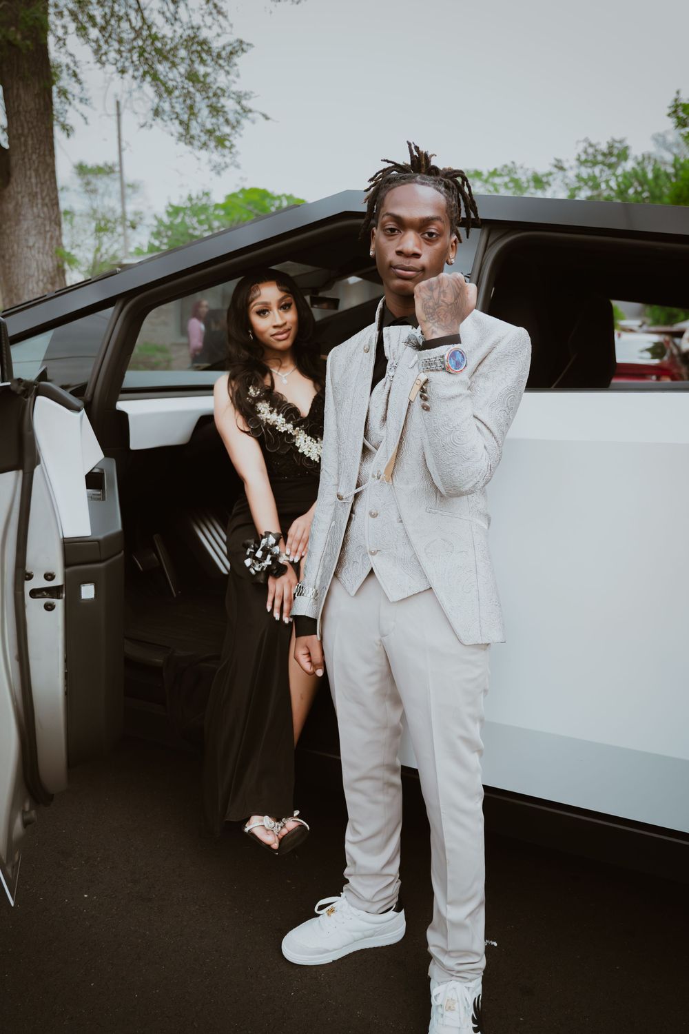 A young couple dressed in prom attire poses beside a white limousine, he in a gray suit and she in a black gown.