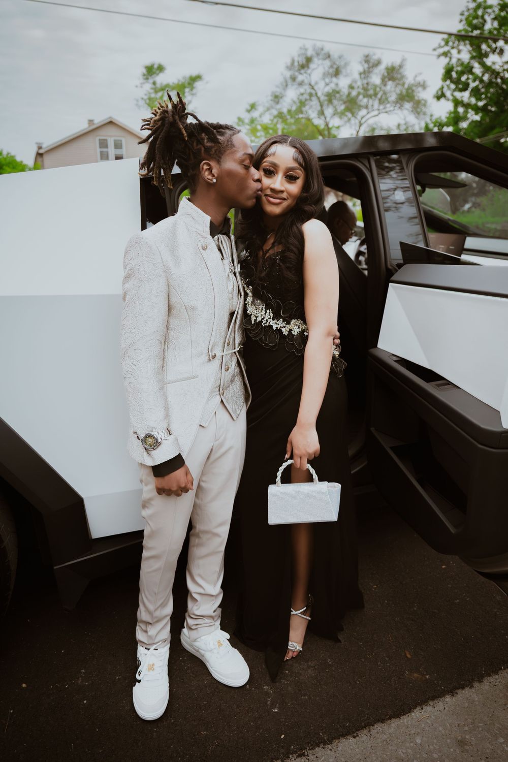 A couple dressed in prom attire, man in white suit kissing woman in black gown, posing beside a white limo.