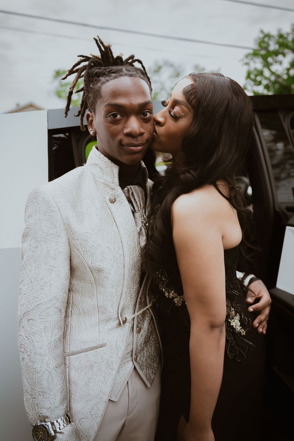 A young couple dressed in elegant prom attire, with a woman kissing a man in a white embroidered suit near a limo.