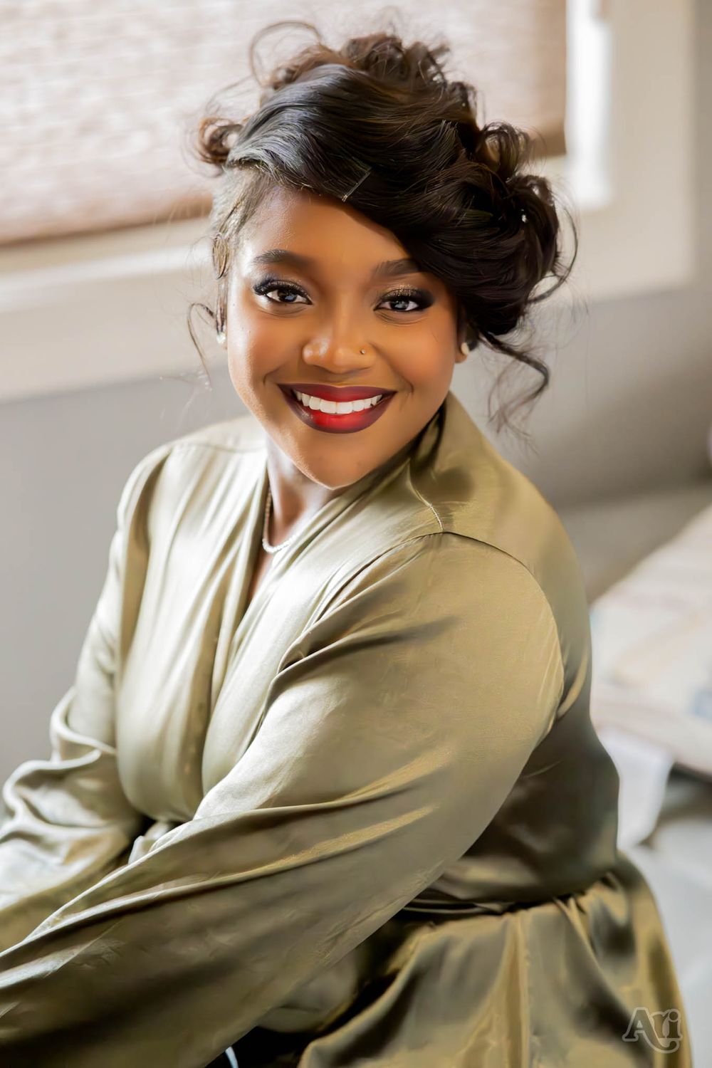 Person in satin olive wrap dress poses elegantly by window with styled hair and makeup.