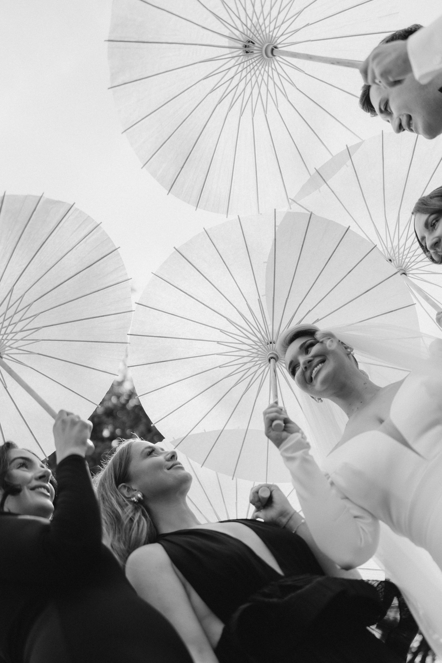 Black and white photo of group holding white parasols viewed from below, smiling upward in elegant attire.