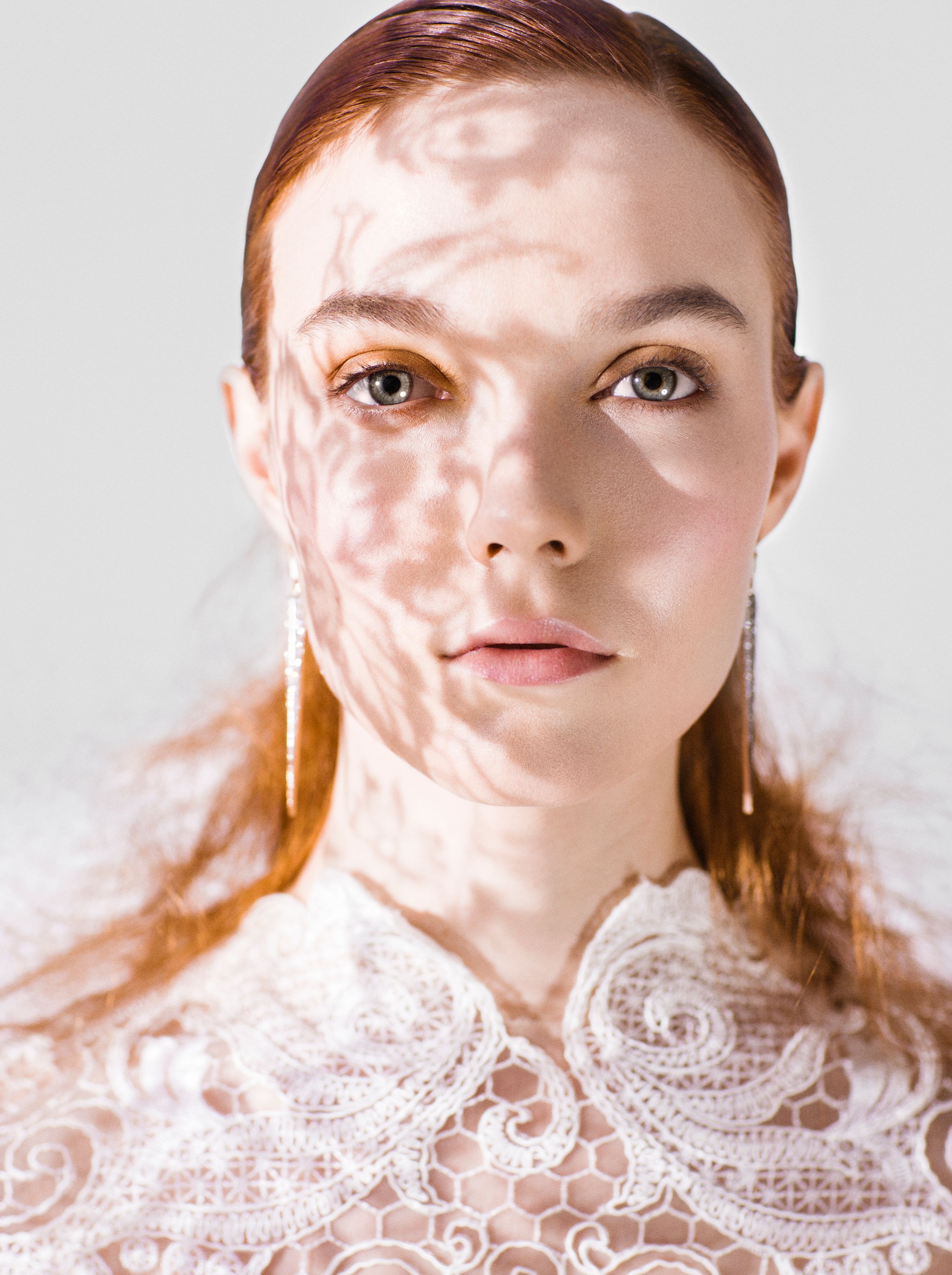 Young redhead woman in white lace top with dappled light shadows on her face, minimalist portrait photography.