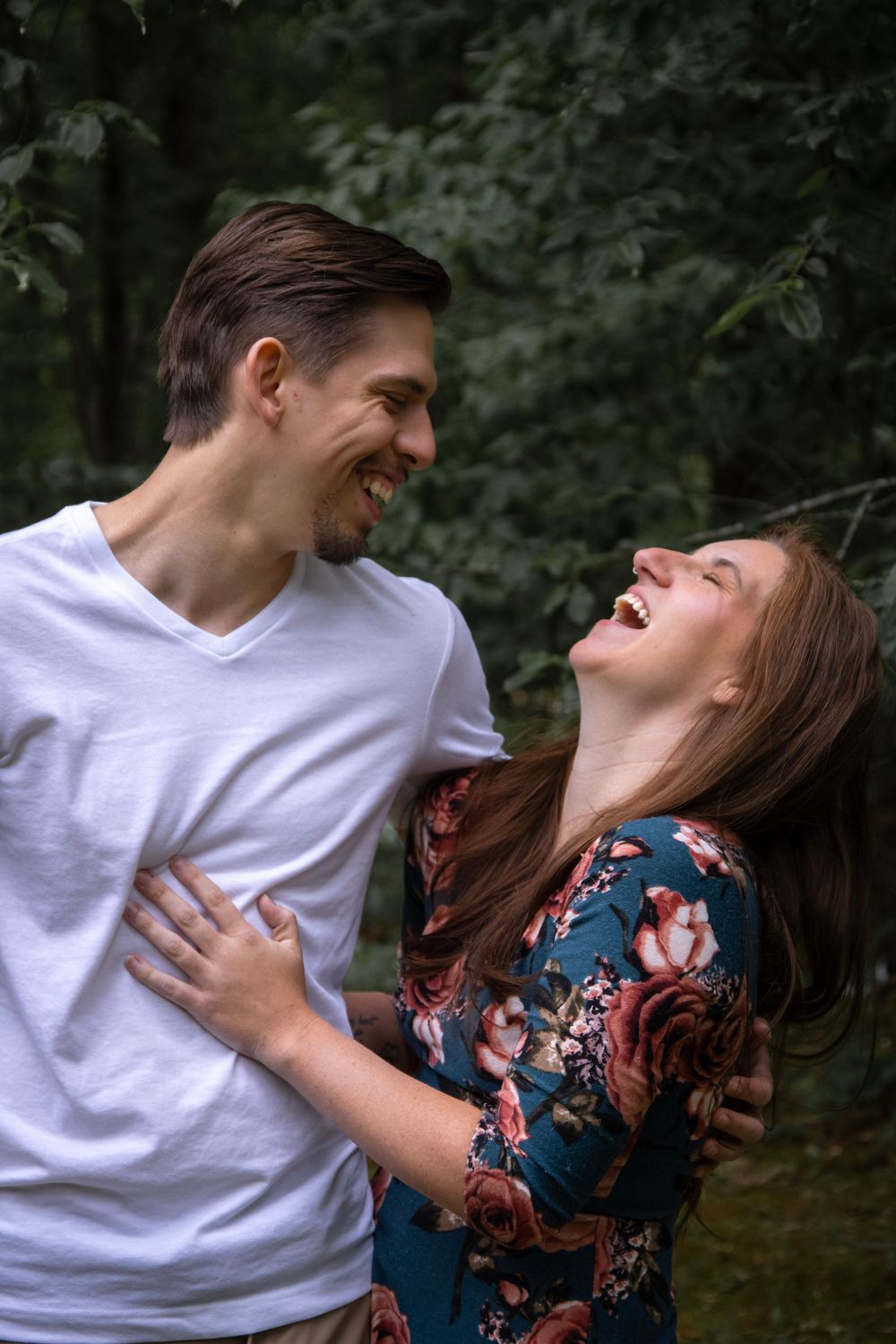 A joyful couple laughing together outdoors, woman in floral dress and man in white t-shirt, surrounded by lush green trees.