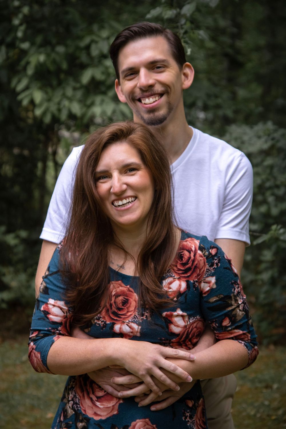 Happy couple embracing outdoors, man in white t-shirt hugging woman in blue floral dress from behind.