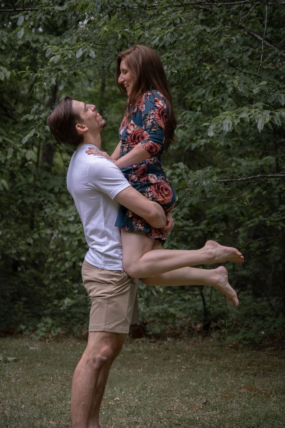 Man lifting smiling woman in floral dress outdoors in lush green park setting.