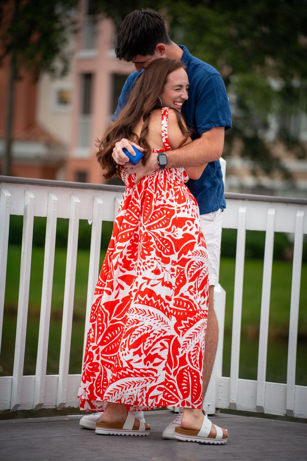 A couple embraces on a white railed bridge, woman in red floral maxi dress, man in blue shirt, sharing a joyful moment.