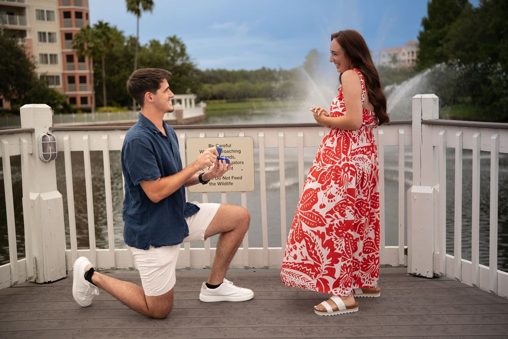 Man proposing on one knee to woman in red floral dress on scenic bridge with fountain backdrop.