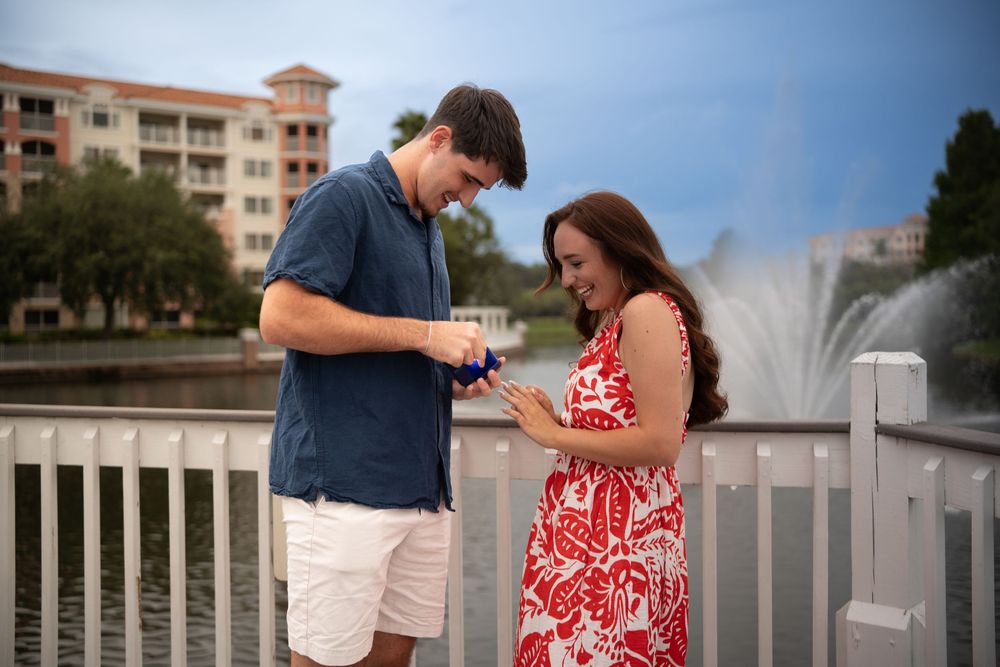 Couple sharing a romantic proposal moment on a bridge by a fountain, man opening a ring box for smiling woman in red dress.
