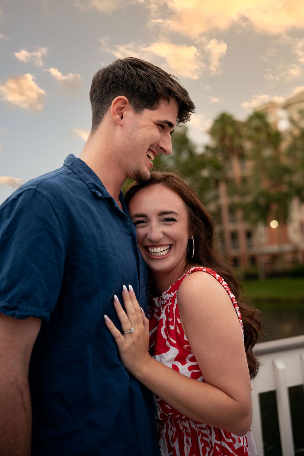 A joyful couple embraces at sunset, woman in red floral dress laughing, man in blue shirt, near a waterfront.