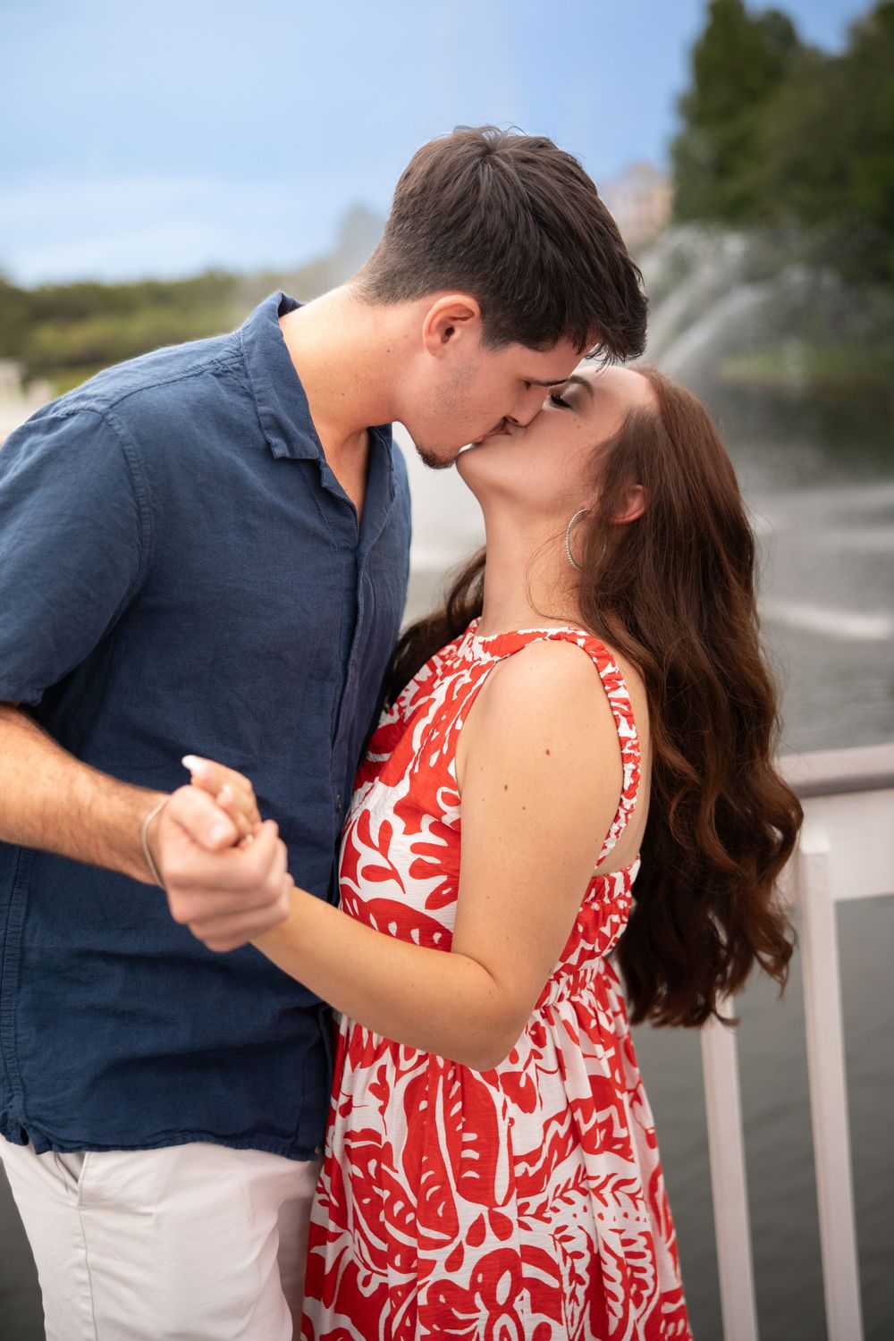 A romantic couple sharing a kiss outdoors, woman in red floral dress holding hands with man in blue shirt near a fountain.