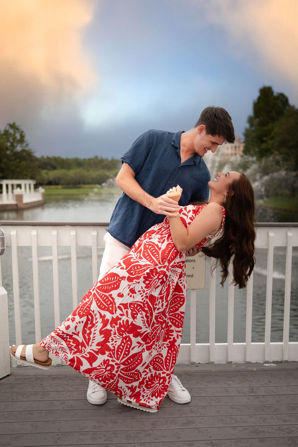 Couple dancing romantically on a waterfront deck at sunset, woman in red floral dress holding ice cream cone.