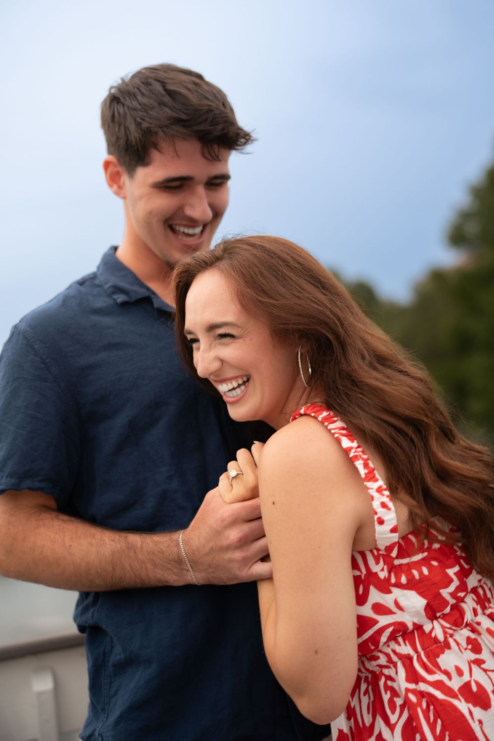 Smiling couple embracing outdoors, woman in red floral dress laughing joyfully as man in navy shirt holds her close.