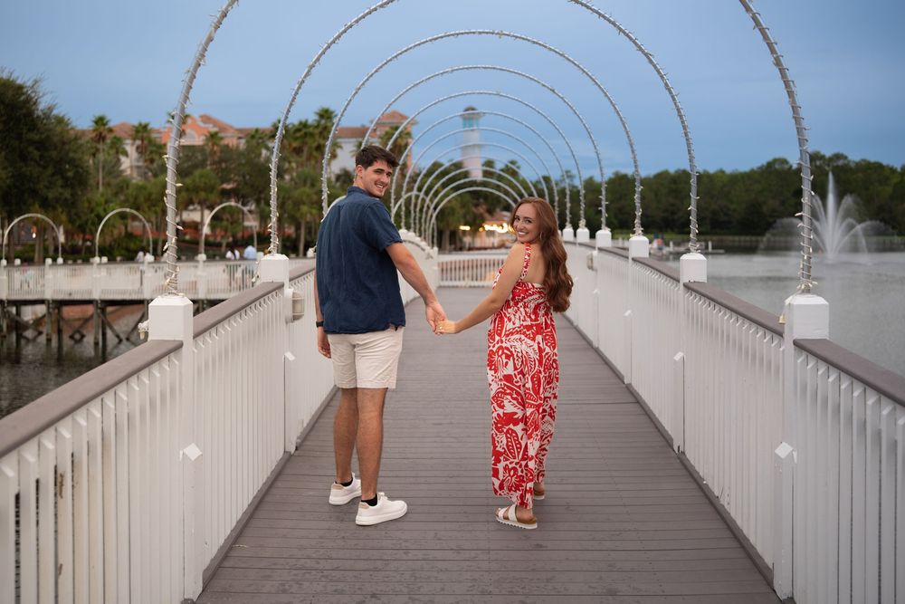 Couple holding hands on a white arched bridge with fountains and trees in the background at dusk.