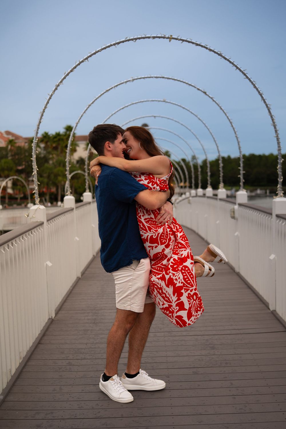 Couple embracing and nearly kissing on a decorative bridge with arch lights, man in blue shirt, woman in red floral dress.