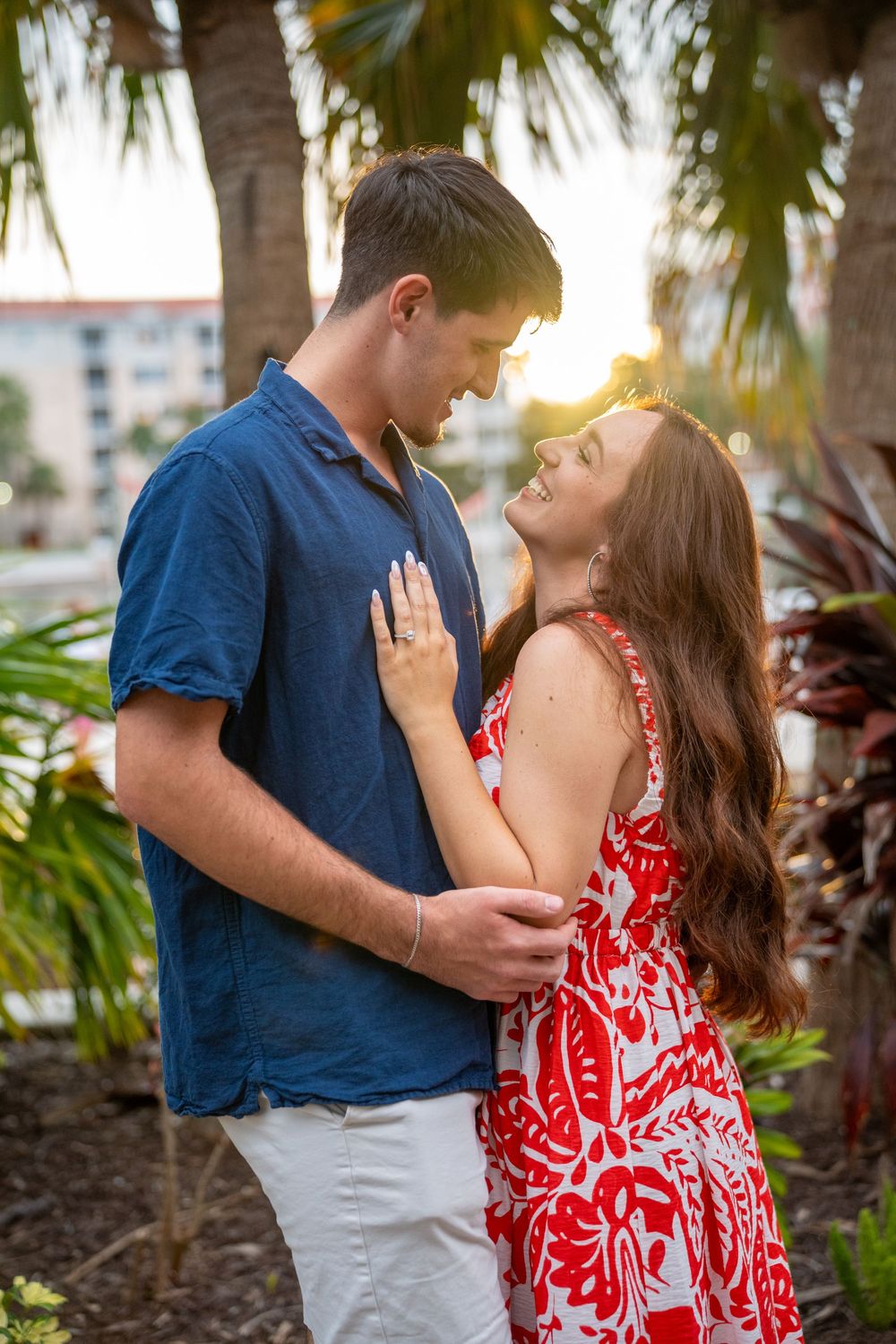 Romantic couple gazing at each other at sunset, woman in red floral dress and man in blue shirt embracing near palm trees.