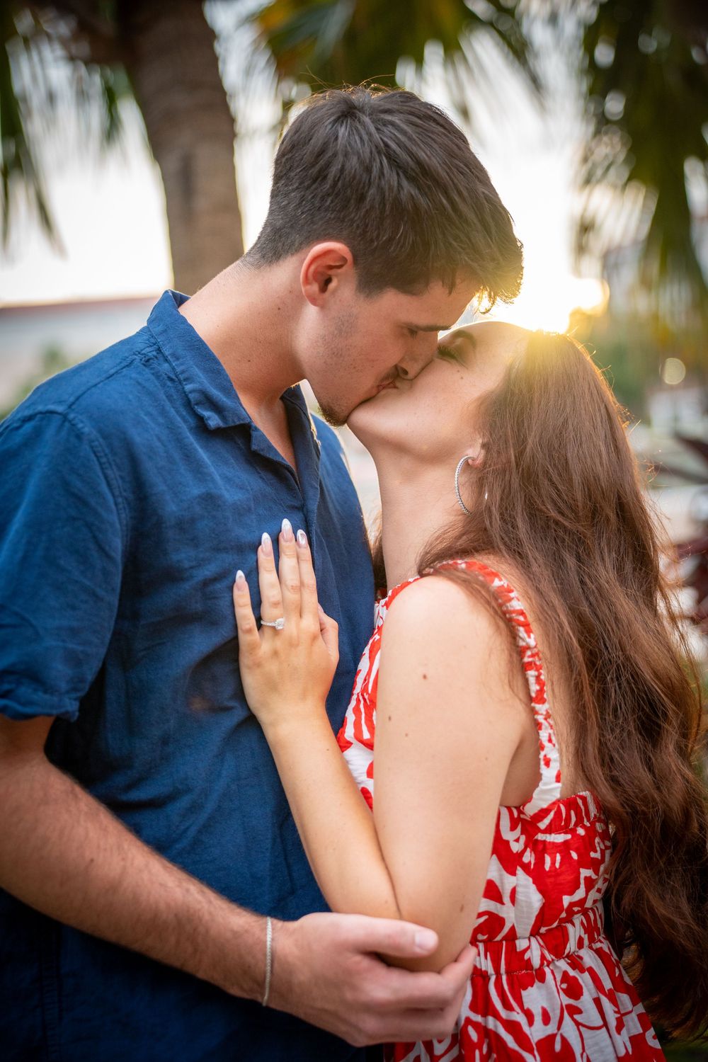 Couple sharing a romantic kiss at sunset near palm trees, woman in red floral dress and man in blue shirt embracing.