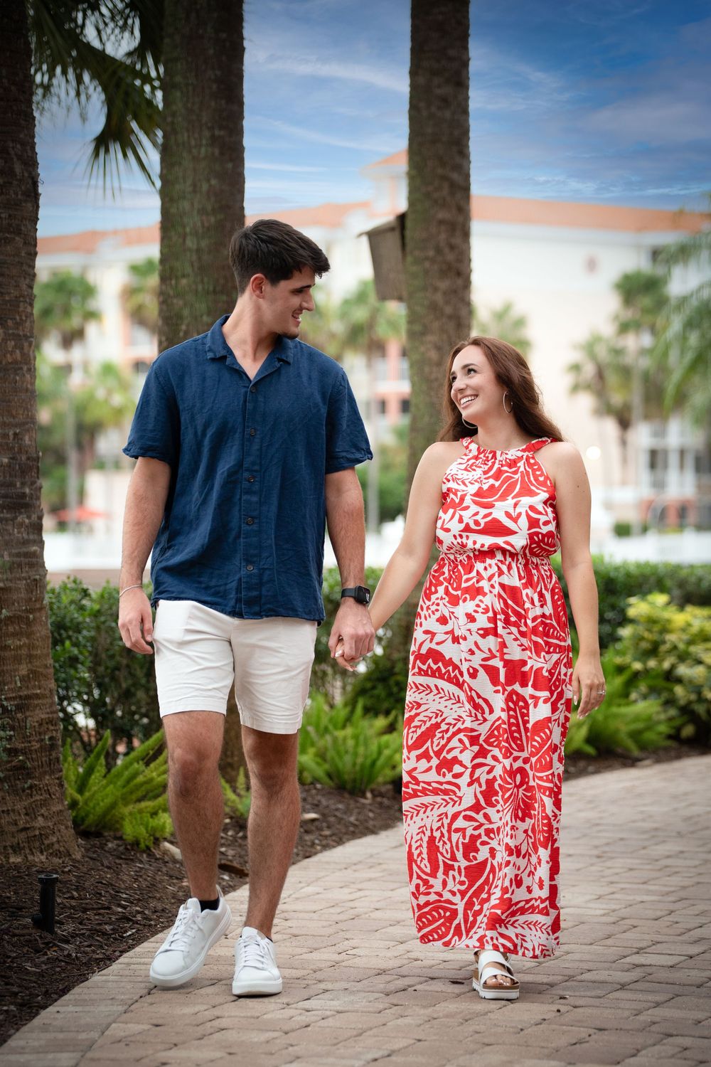 A couple holds hands and walks along a palm-lined path at a tropical resort, dressed in summer attire.
