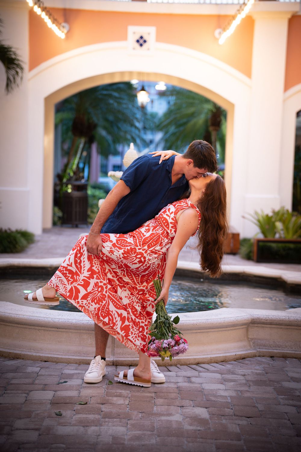 Romantic couple sharing a dip kiss by a fountain, woman in red floral dress holding pink flowers, elegant courtyard setting.