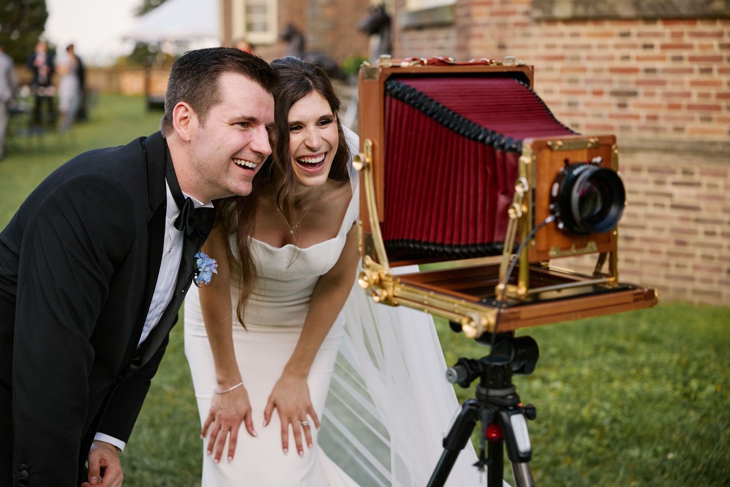 Bride and groom laughing together beside a vintage large format camera on a tripod at an outdoor wedding reception.