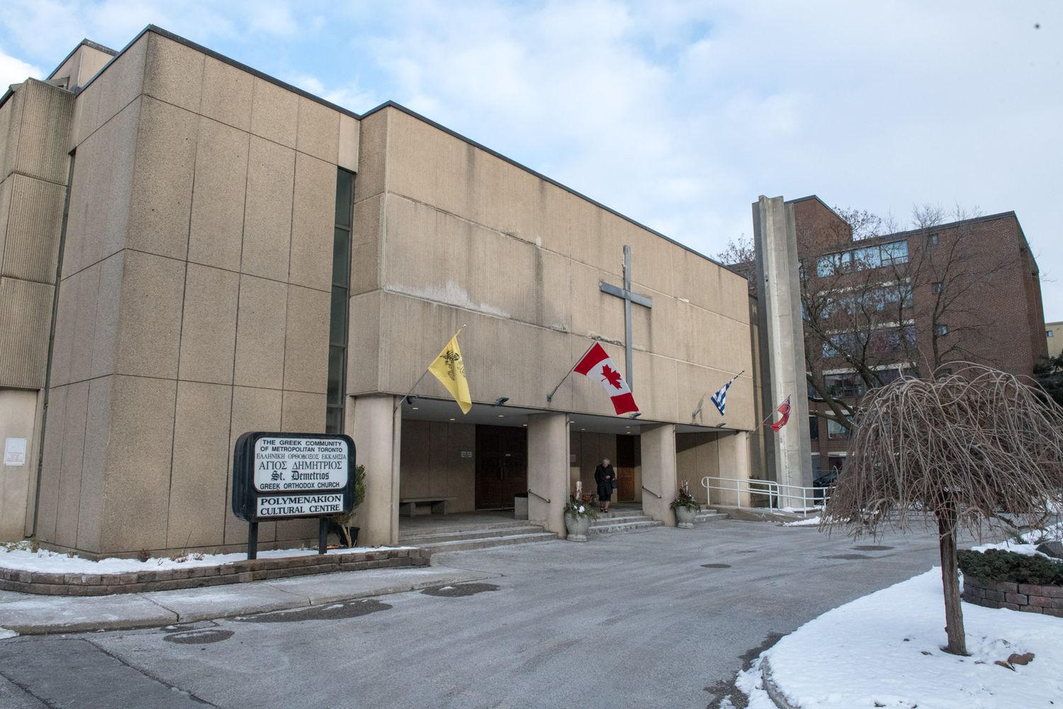 Exterior of a concrete church building with Canadian flags and a cross in winter snow.