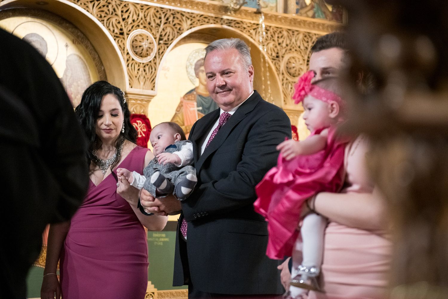 Man in suit holds baby boy in gray outfit surrounded by guests at an Orthodox church baptism.