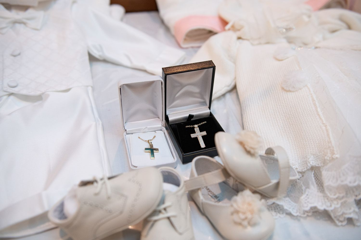 Silver cross necklaces in jewelry boxes displayed with white christening shoes and clothing.