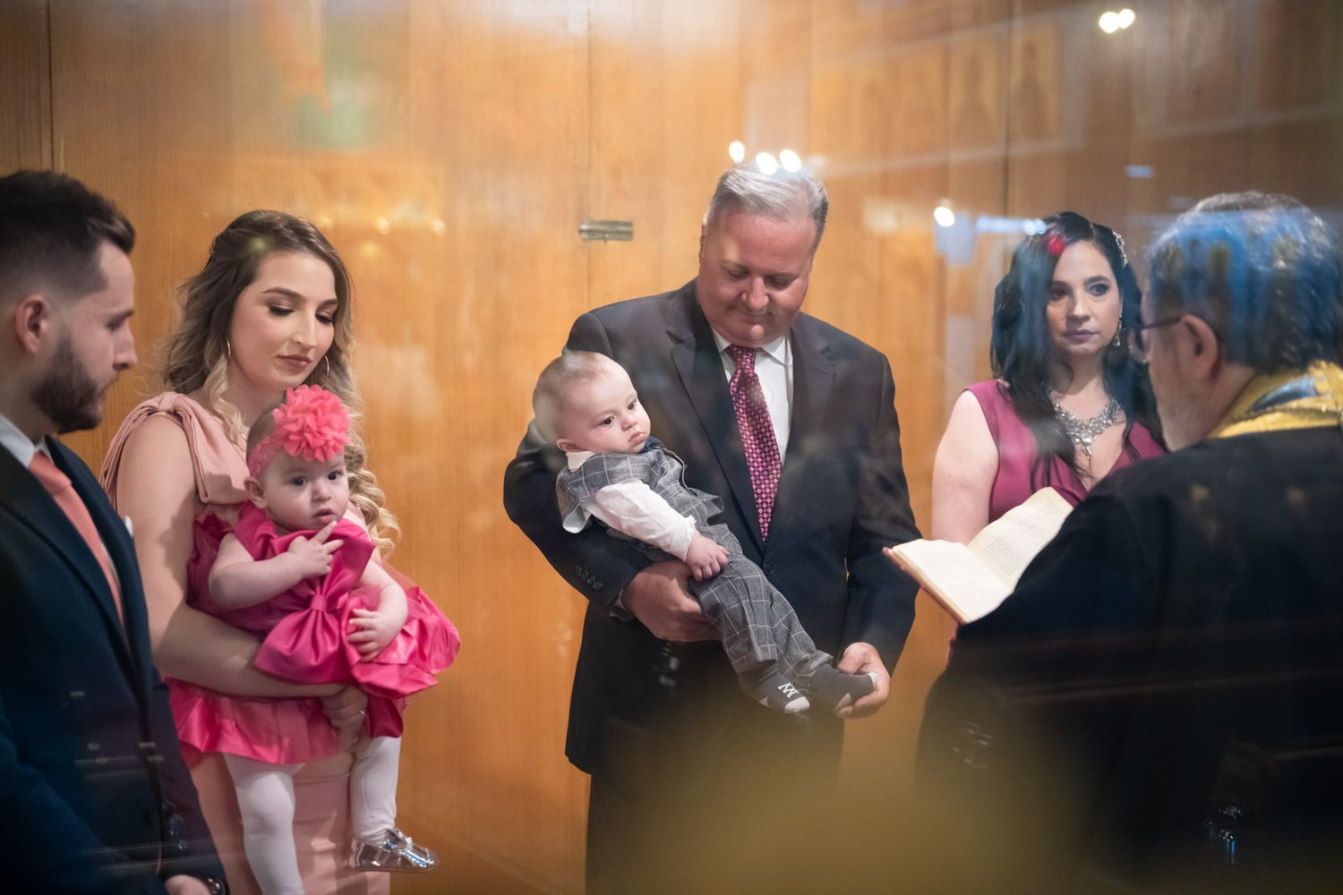 Priest performing a christening ceremony holding a baby with family gathered around.