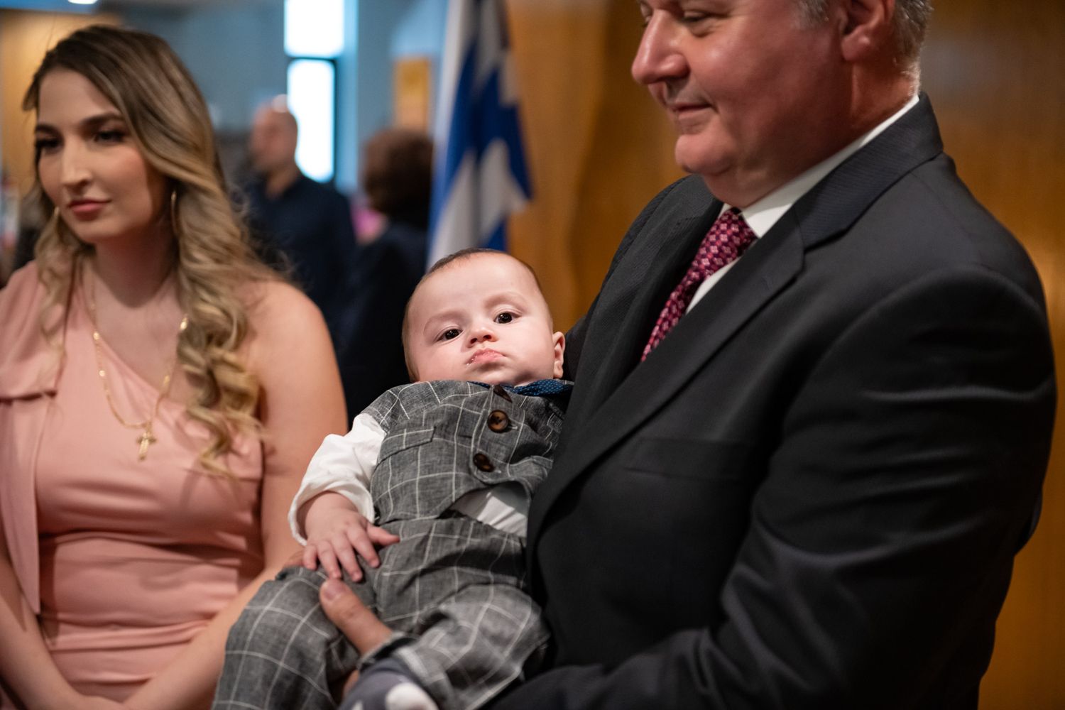 Older man in a suit holding a baby boy in a gray outfit at a christening event.