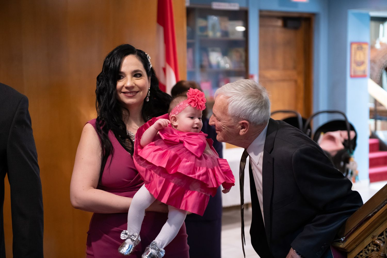 Woman in pink dress holds baby girl in pink outfit while elderly man leans in to kiss her at a christening.