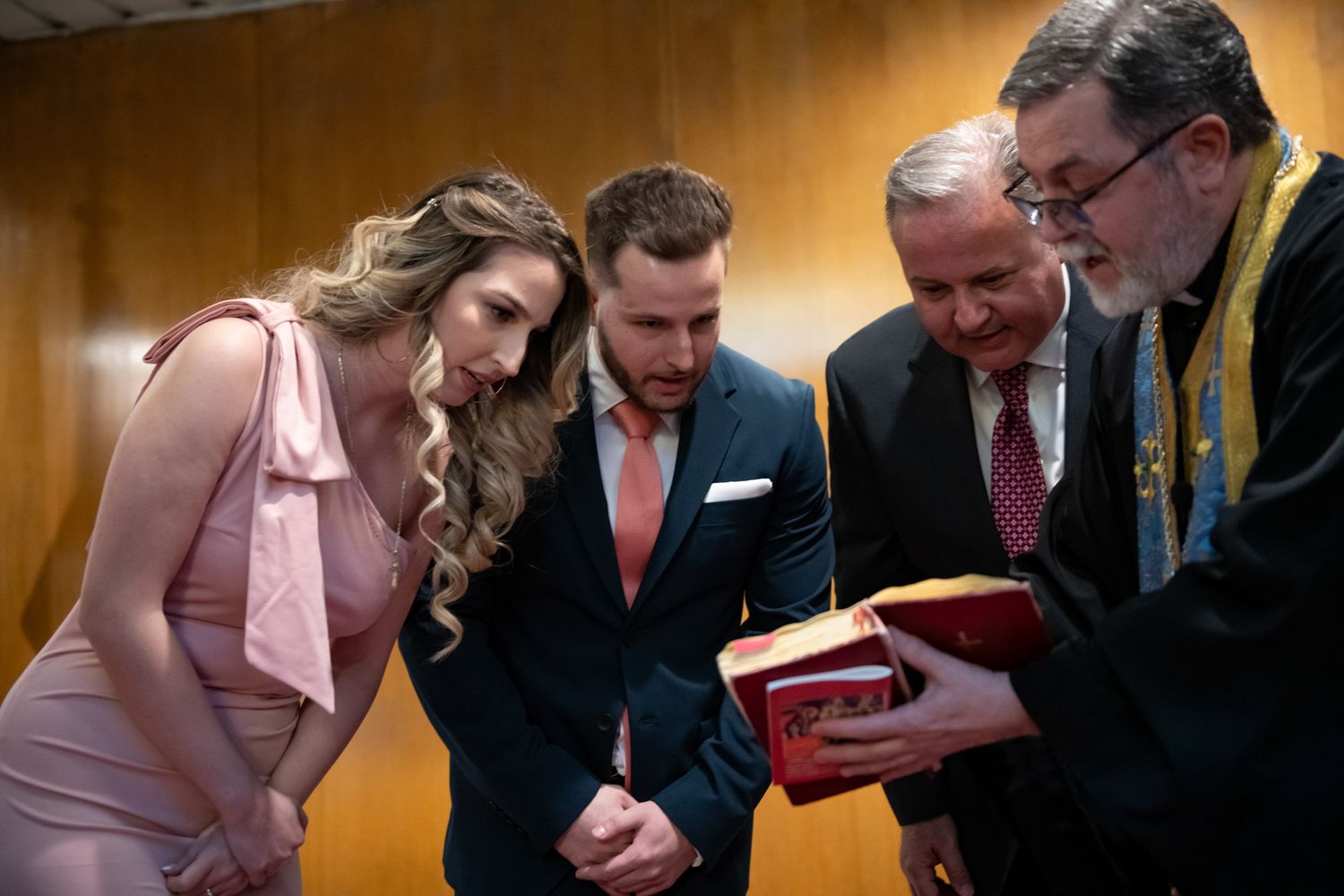 Couple in formal attire reviews religious text with two men including a priest at a baptism ceremony.