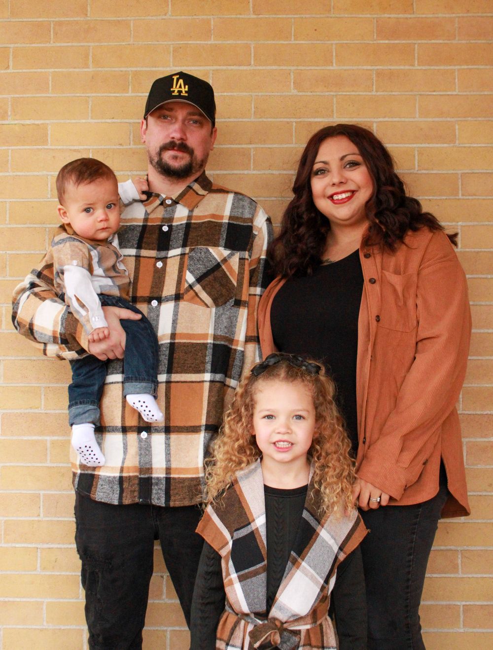Family of four posing against a brick wall, parents with two young children wearing coordinating plaid outfits.