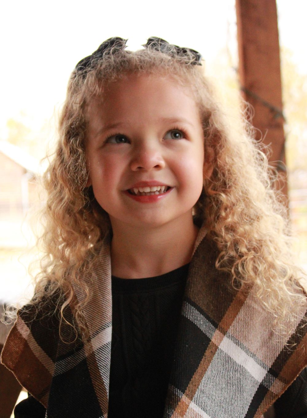 Curly-haired little girl in plaid cape smiles joyfully outdoors in fall setting.