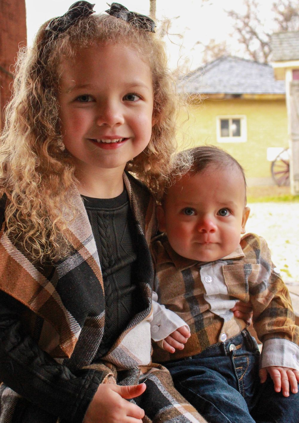 Curly-haired girl in plaid vest poses with baby sibling in matching plaid shirt for fall family photos.