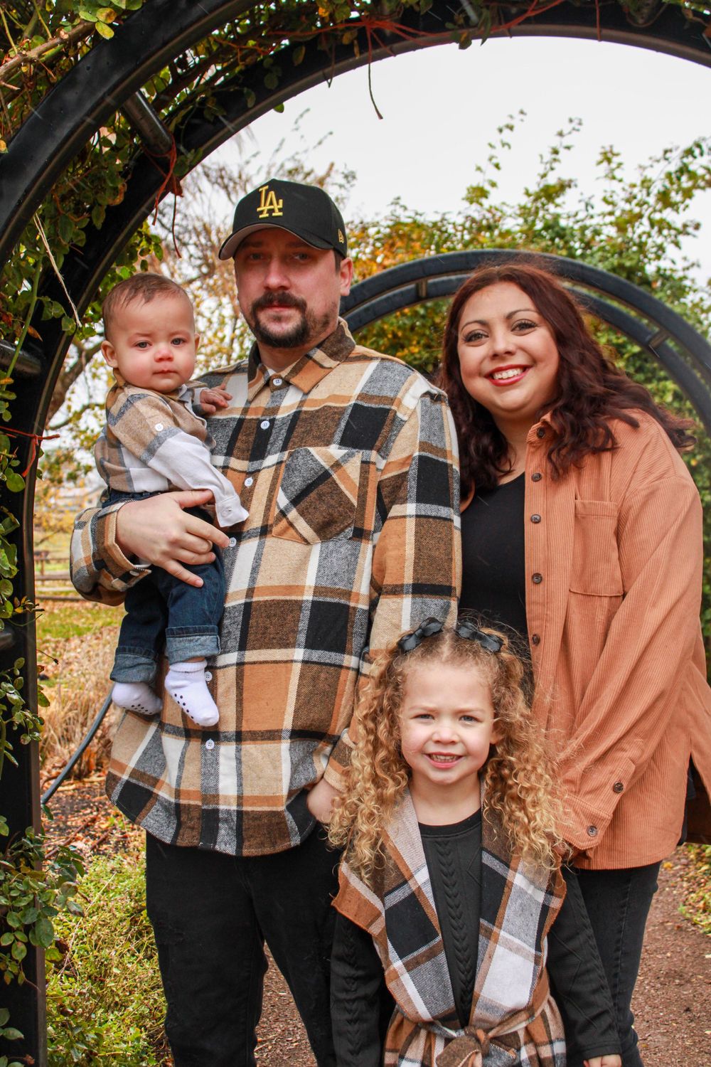 Happy family of four posing under garden arch in autumn, father holding baby, young girl in front.