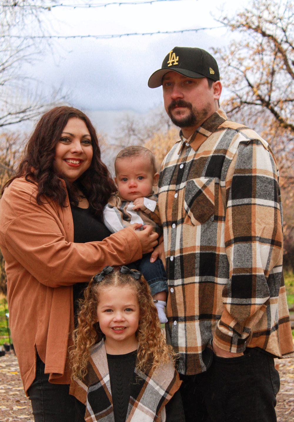Family of four posing outdoors in winter, parents holding infant baby with young curly-haired girl.