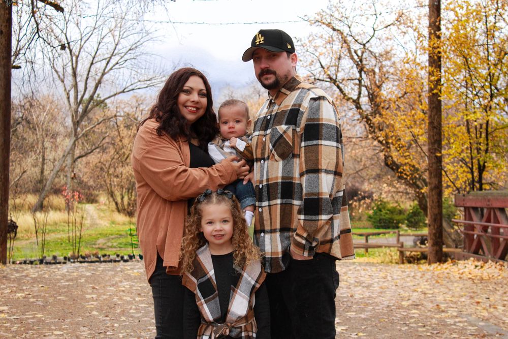 Family of four posing outdoors in autumn, parents holding young children among colorful fall foliage.
