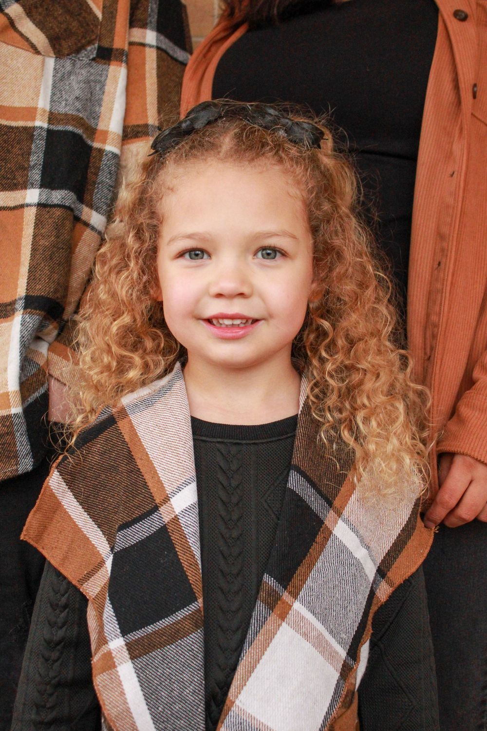 Young girl with curly blonde hair wearing a plaid vest smiles at camera during fall family photo session.