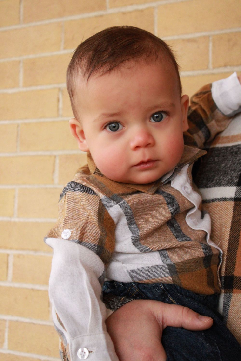 Adorable baby boy with blue eyes wearing a plaid flannel shirt being held by an adult.