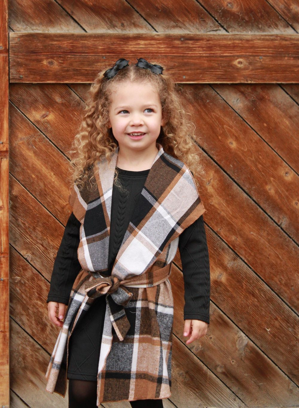 Young girl in plaid vest with brown belt poses in front of rustic wooden barn door.