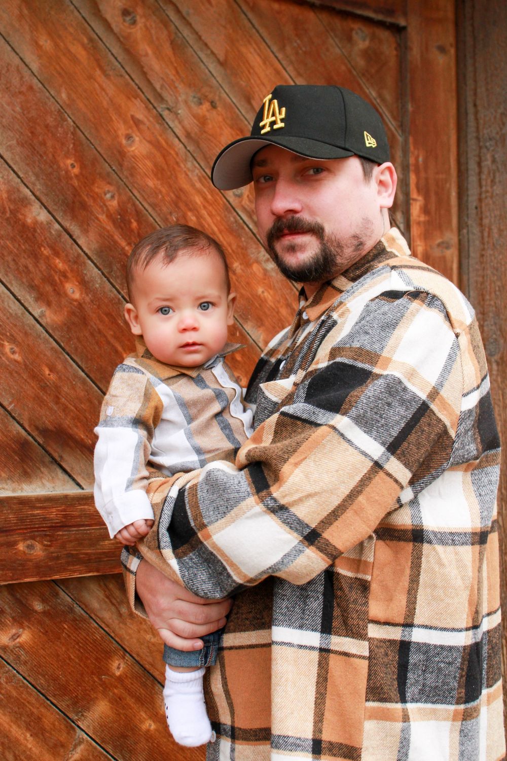 Father in LA cap and plaid shirt holding his infant baby in front of rustic wooden door.