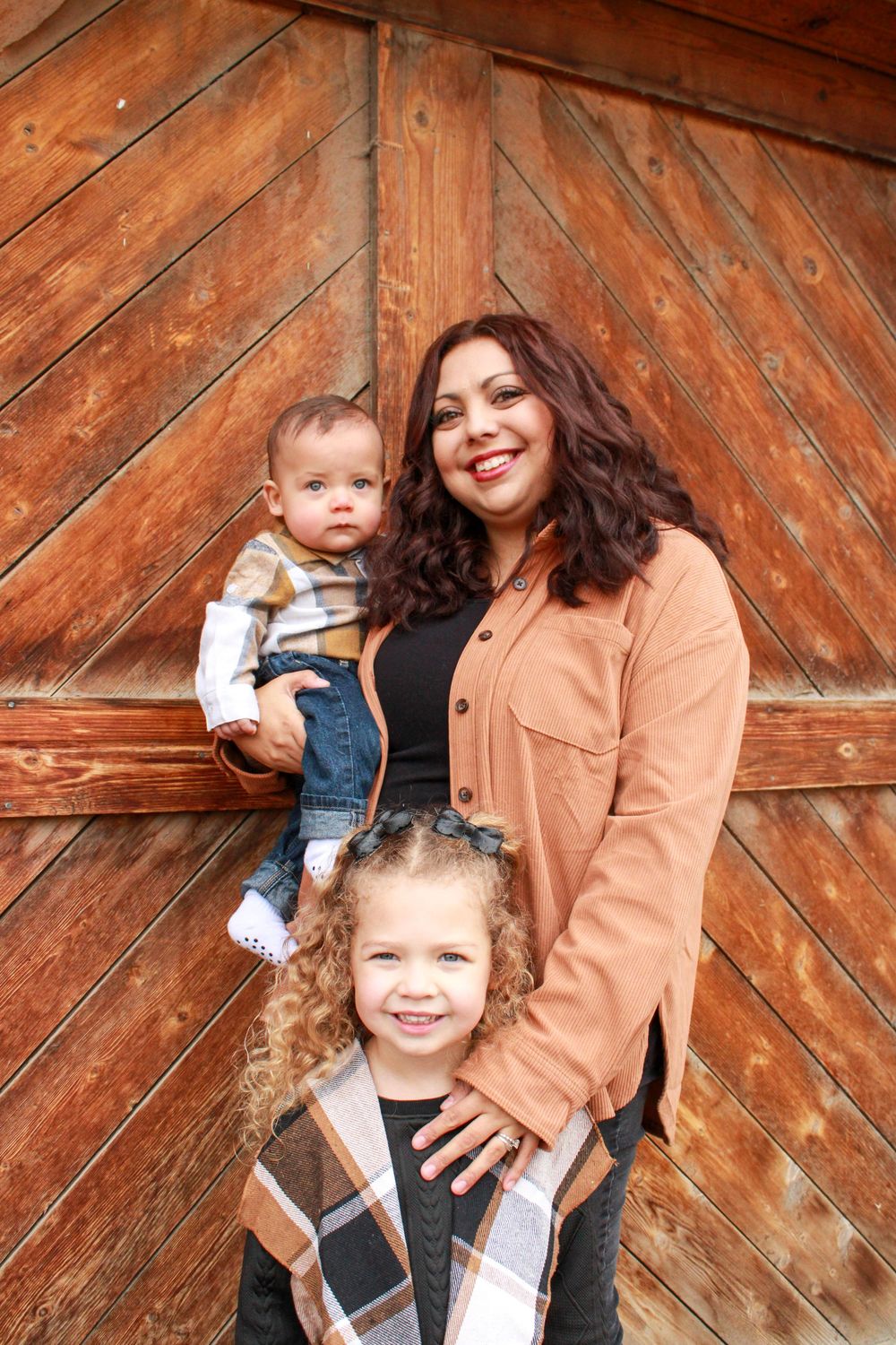 A smiling woman in a rust jacket holds a baby while a curly-haired toddler stands in front, posed by a wooden barn door.