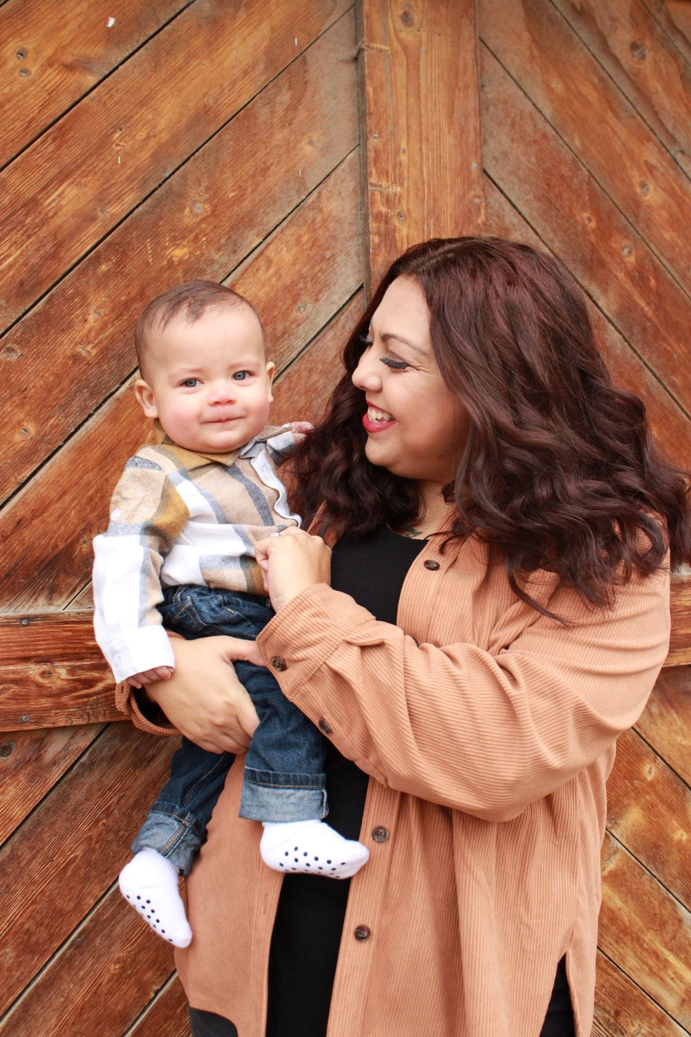 Smiling mother in rust orange shirt holds her baby boy in plaid outfit against rustic wooden backdrop.