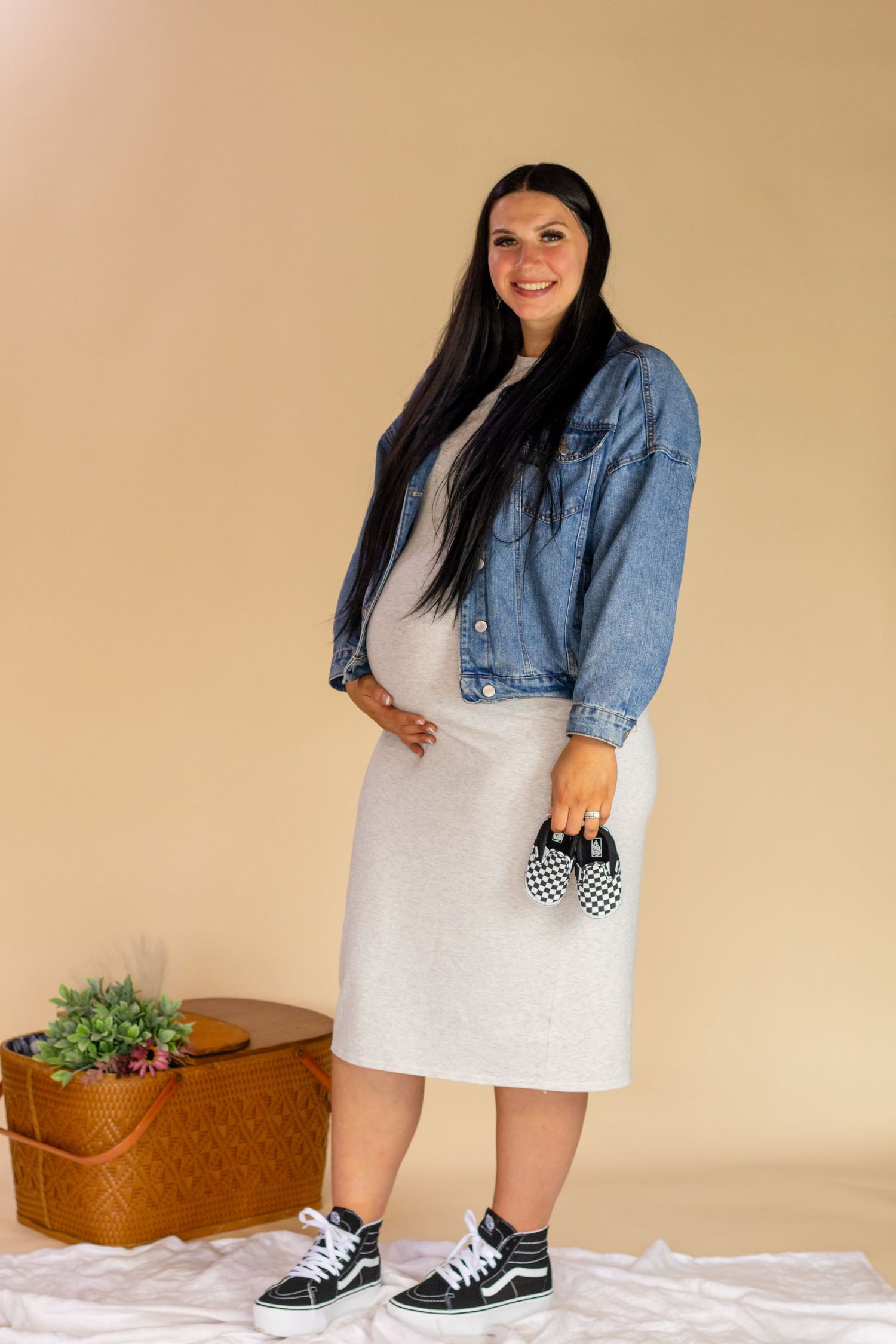 Smiling pregnant woman in denim jacket and gray dress holding checkered baby shoes in roy wa studio with pierce county photographer.