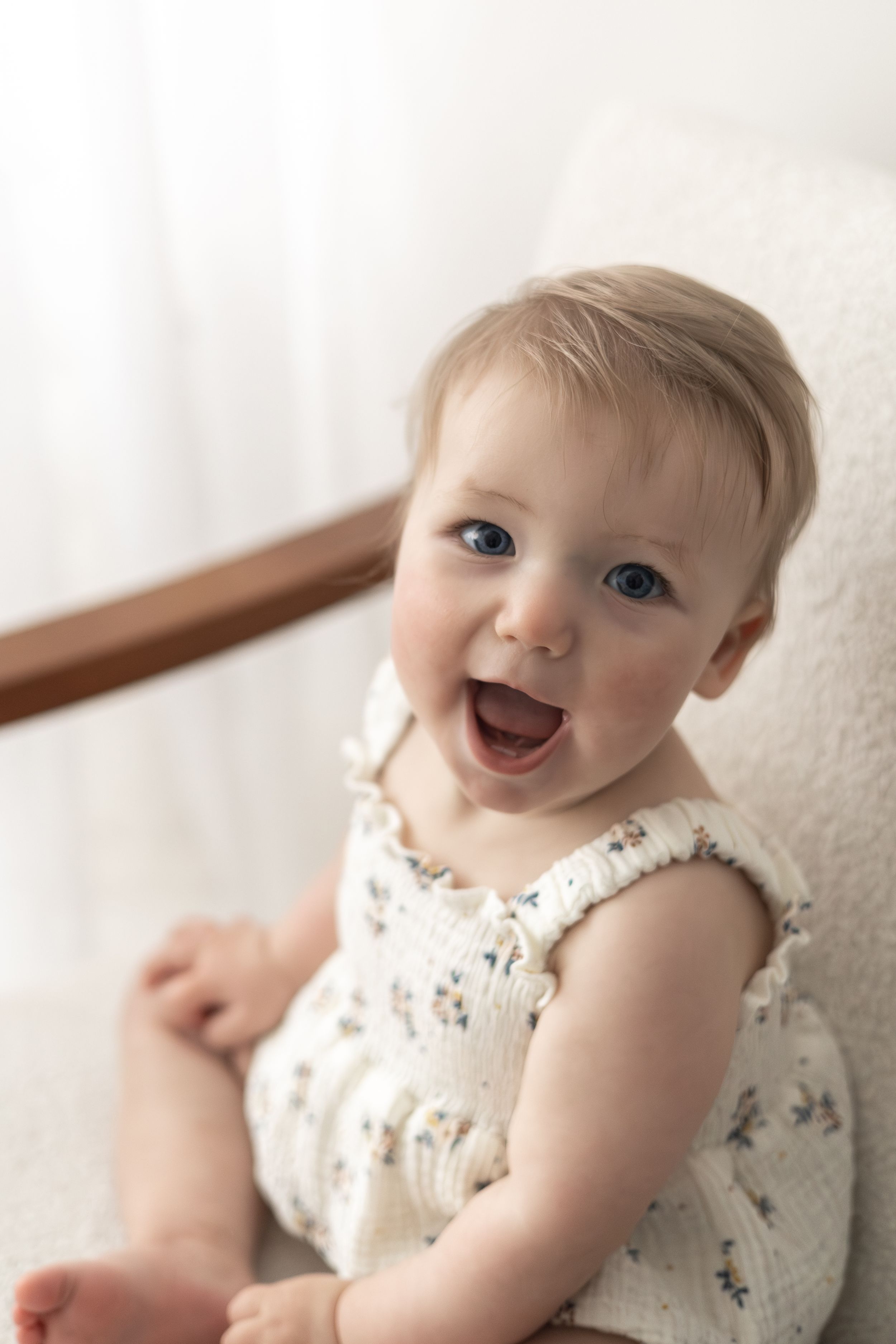 Smiling baby with blue eyes wearing a white floral romper sitting on a white chair birthday photography Nottingham baby turning one