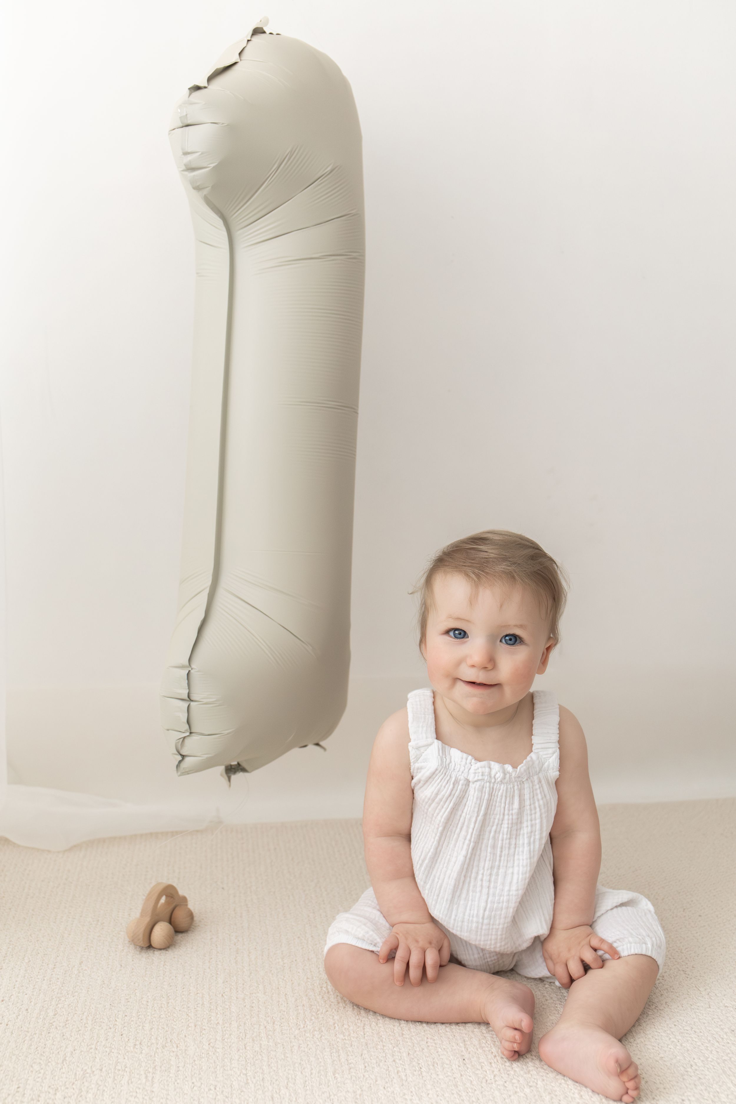 Baby girl in white romper sits smiling beside a large number 1 balloon, celebrating her first birthday photography Nottingham