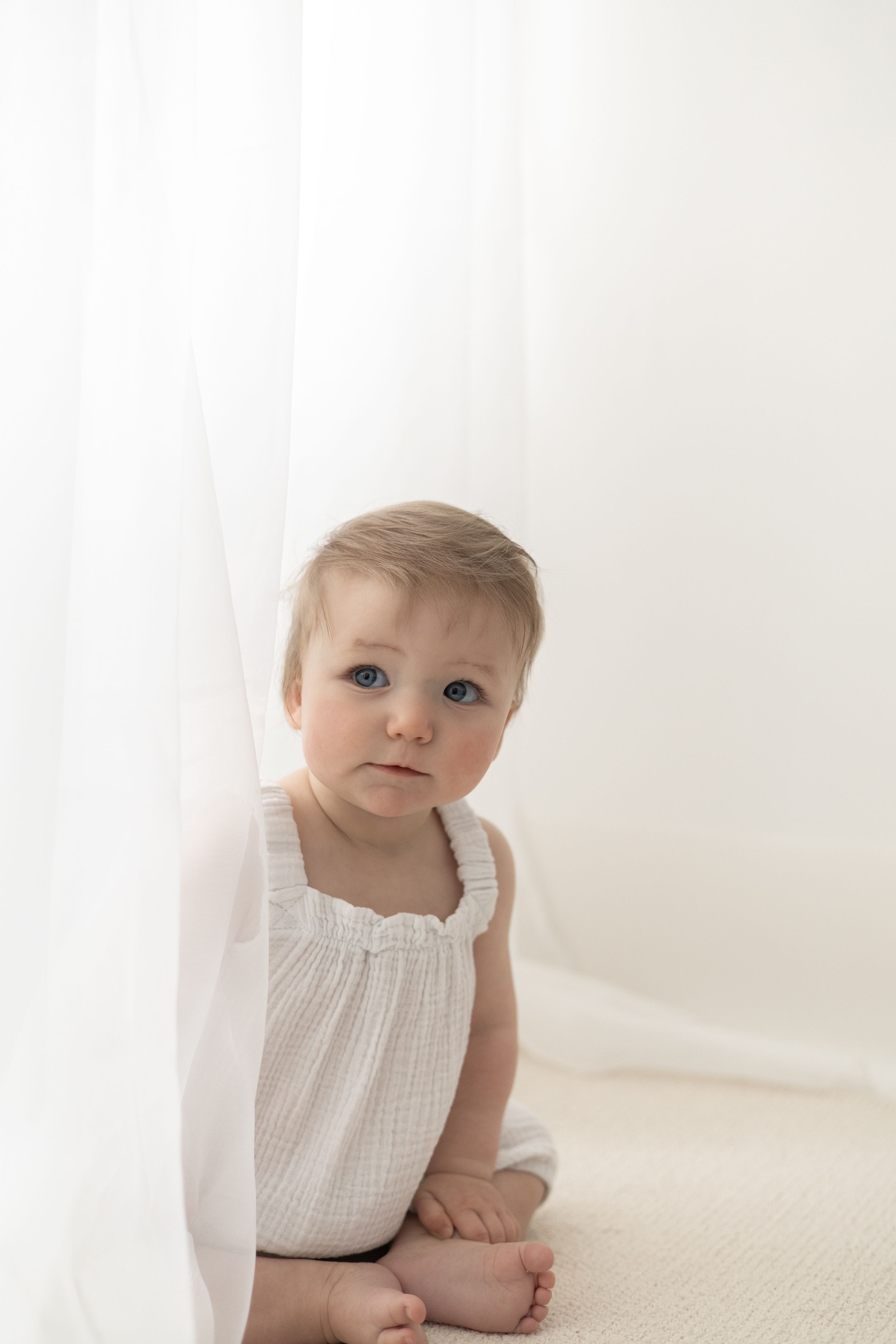 Adorable baby girl in white romper sitting on carpet beside sheer white curtain, natural birthday photography Nottingham baby turning one