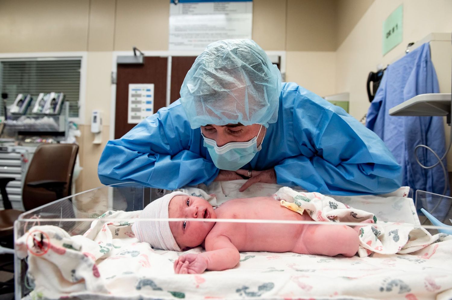 new dad in protective equipment examines newborn baby in hospital bassinet.