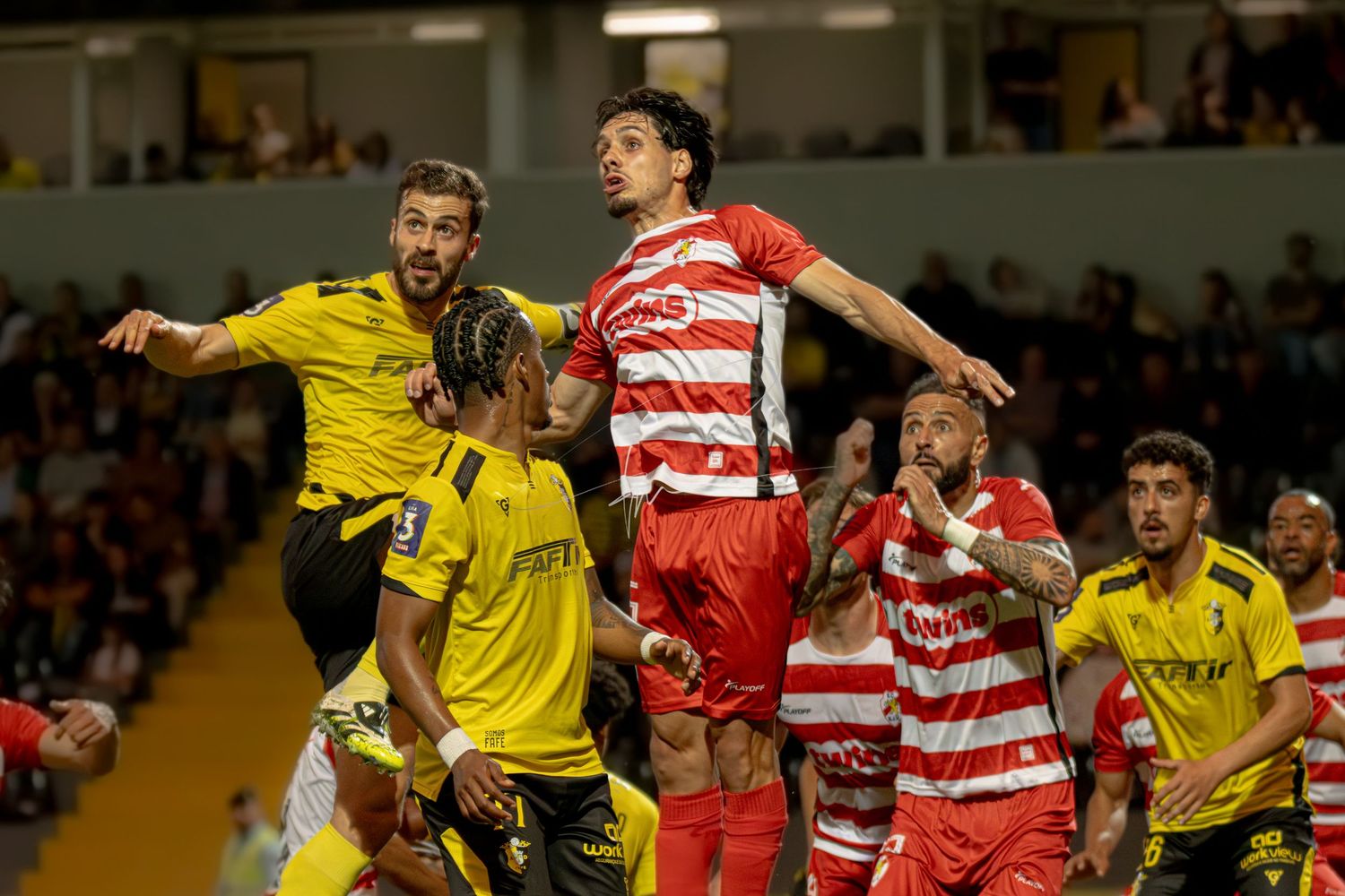 Soccer players in red and yellow jerseys compete for a header during an intense football match at night.