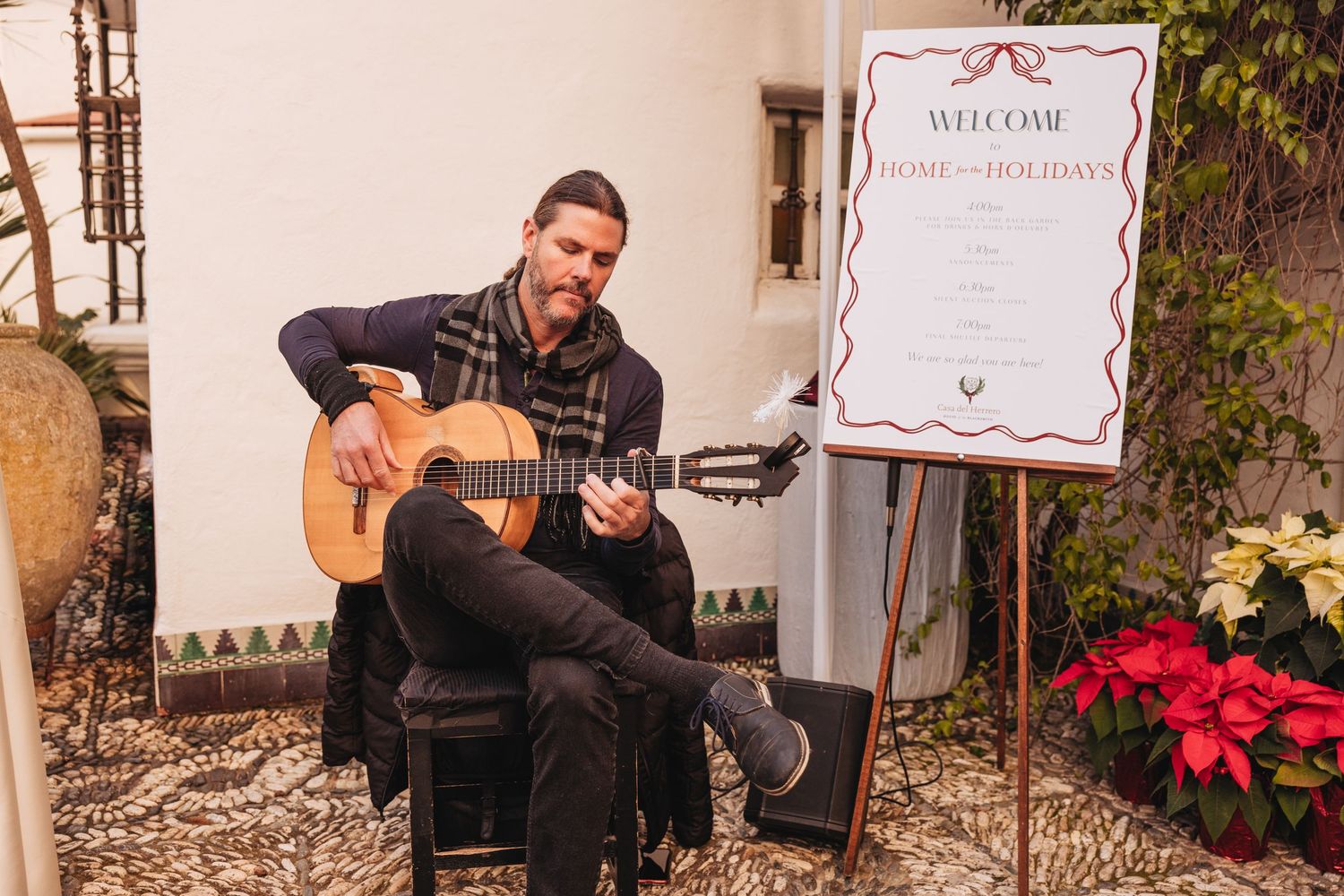 A man playing acoustic guitar at a holiday event beside a Welcome Home for the Holidays sign with poinsettias.